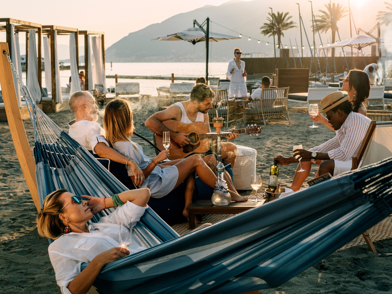 Group of friends relaxing on the beach at sunset in Porto Montenegro, with one person playing guitar and others enjoying drinks.