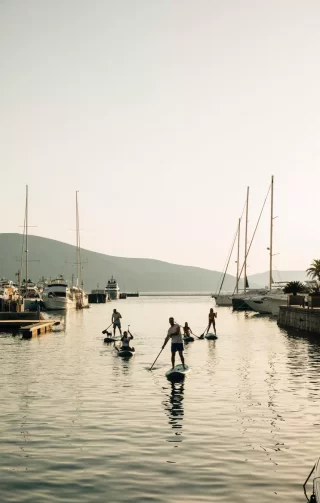 Group of people paddleboarding at sunset in Porto Montenegro, navigating through the marina with sailboats and yachts docked nearby, surrounded by calm waters and mountains in the background.