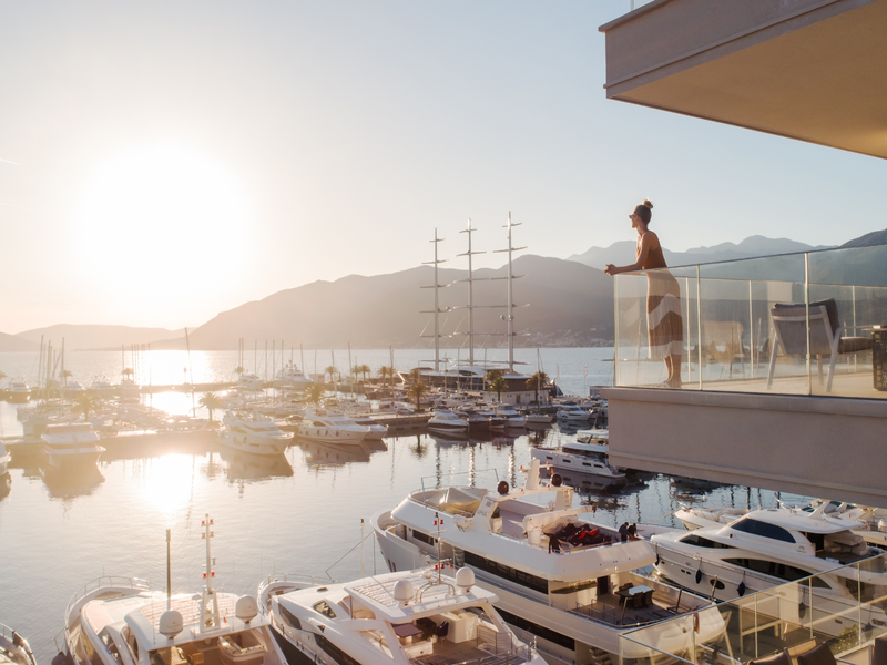 Woman standing on a luxurious balcony overlooking a marina filled with yachts at sunset, with mountains in the background and the sun setting over the water.Woman standing on a luxurious balcony overlooking a marina filled with yachts at sunset, with mountains in the background and the sun setting over the water.