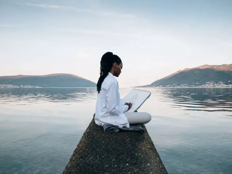 Woman sketching on a notepad while sitting on a pier overlooking a calm lake with mountains in the background.