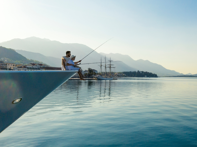 Father and son fishing from the bow of a yacht in Porto Montenegro, with mountains and calm water in the background.