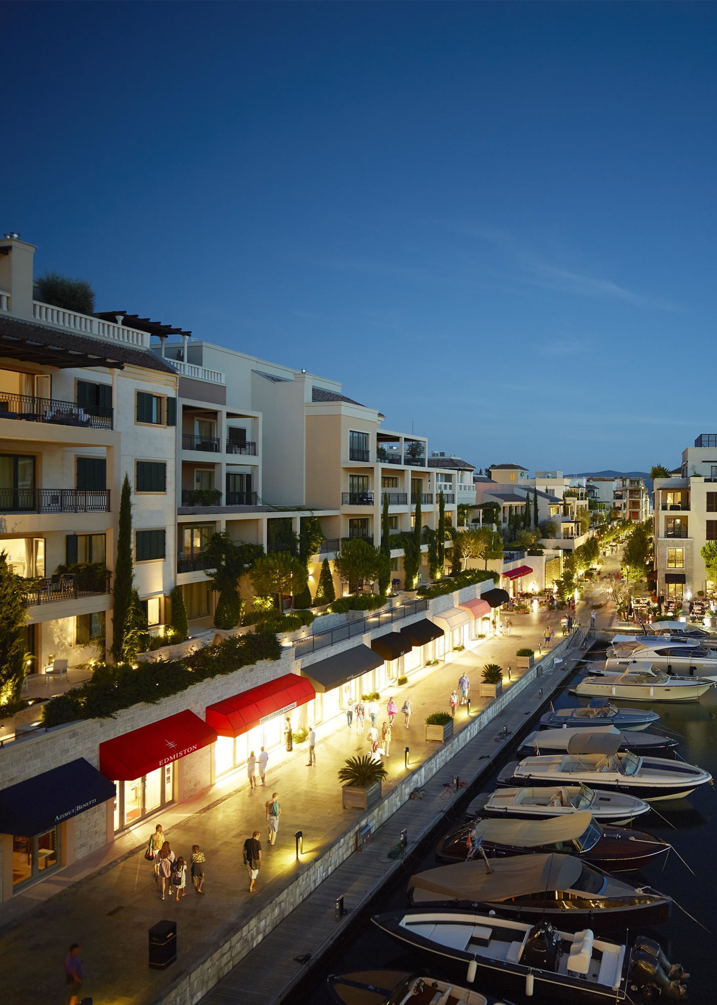 A bustling waterfront promenade at dusk, lined with modern residential buildings and boats moored along the docks, illuminated by streetlights and vibrant shop fronts, creating a lively evening atmosphere.