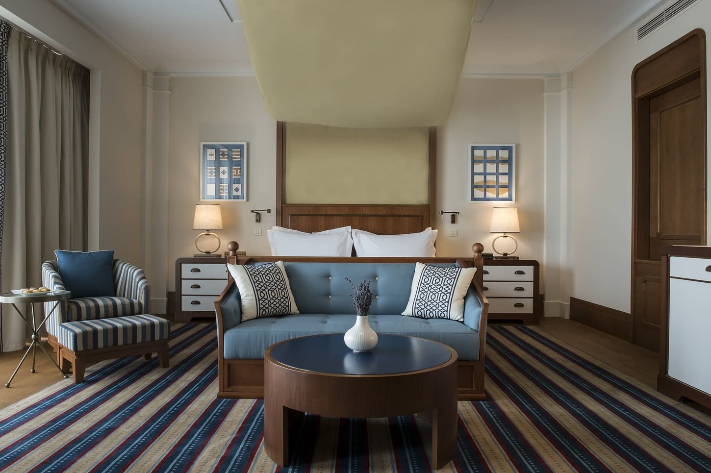 A modern and elegantly decorated hotel room featuring a wooden bed with plush bedding, striped upholstered chairs, a round coffee table, and a colorful striped rug.