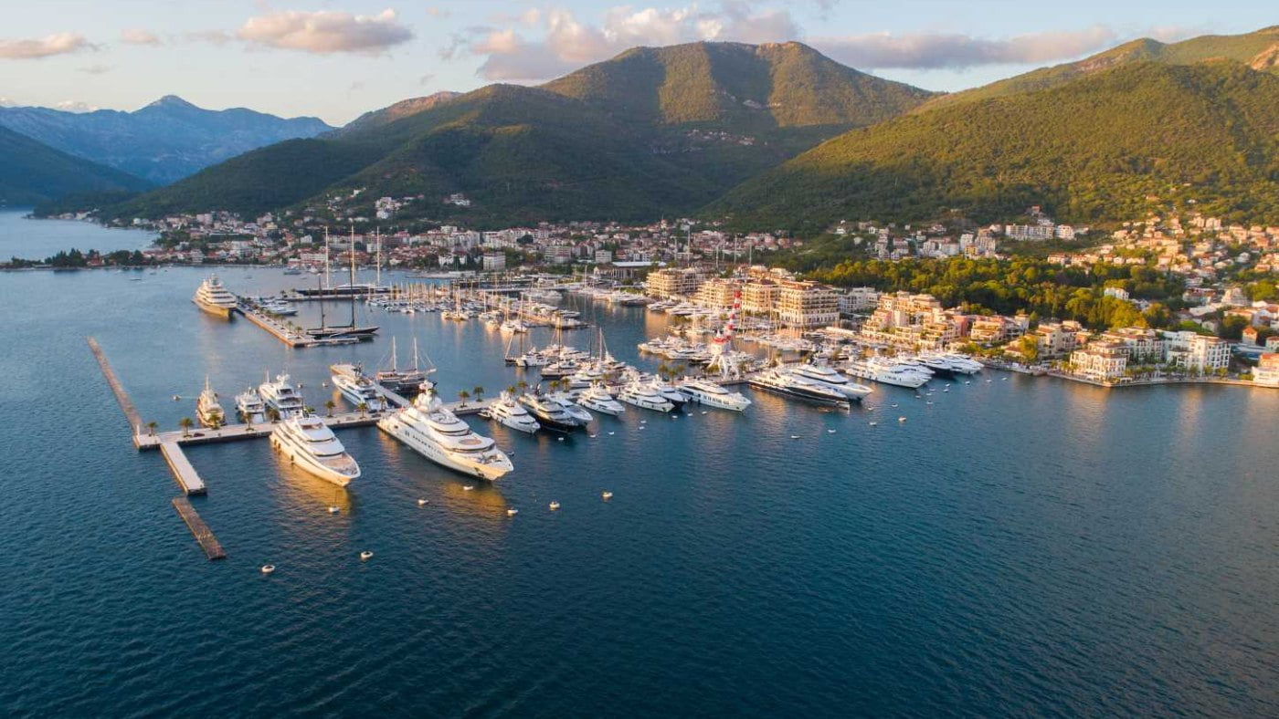 An aerial view of a coastal town at sunset, with a marina filled with docked yachts and a mountainous backdrop.