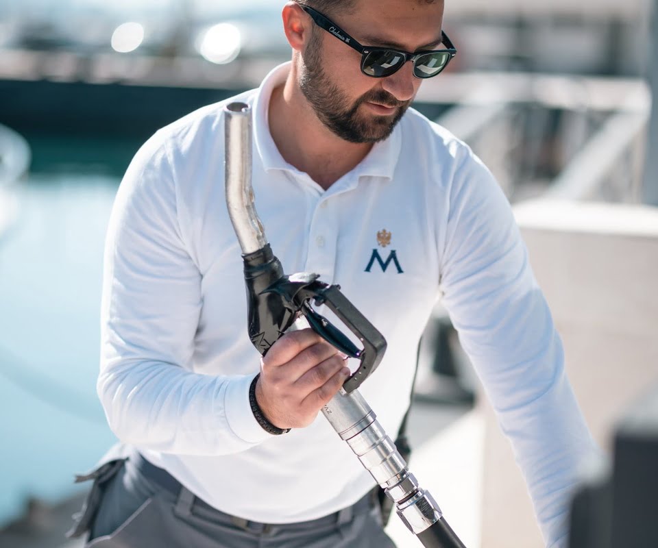 A man in sunglasses and a white polo shirt holds a yacht fuel pump nozzle, likely preparing to refuel a vessel.