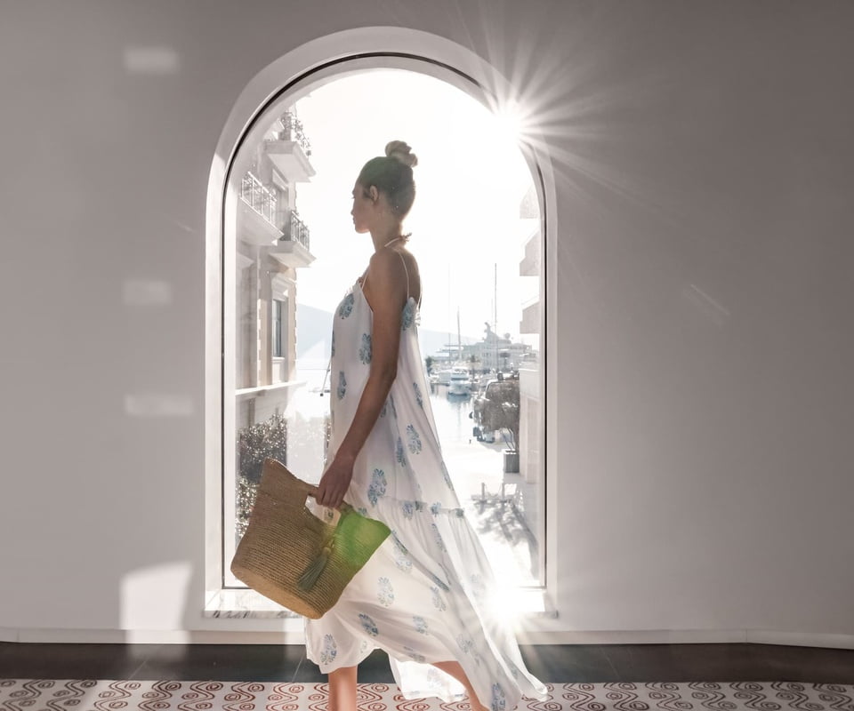 A woman in a flowing white dress with blue patterns stands in a sunlit arched doorway holding a straw tote bag.