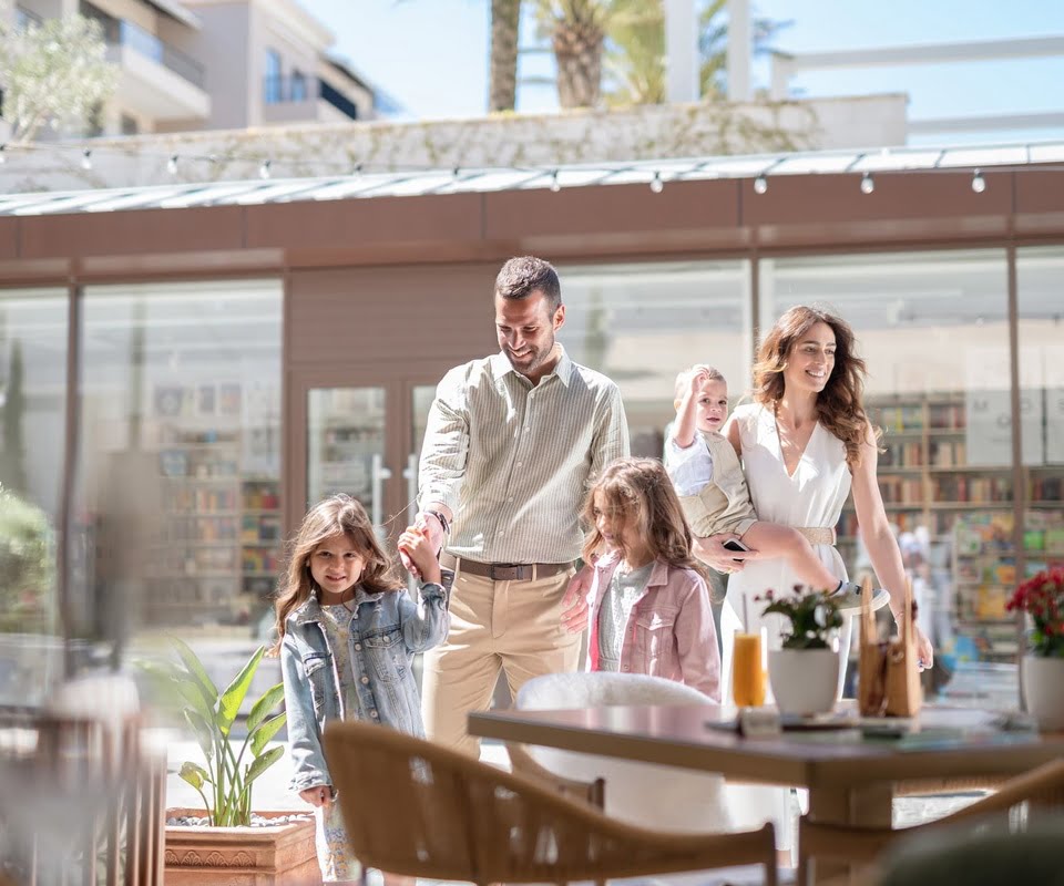 A family is walking through a sunny outdoor cafe area, with the parents holding hands with their two young daughters, and the mother carrying a toddler.
