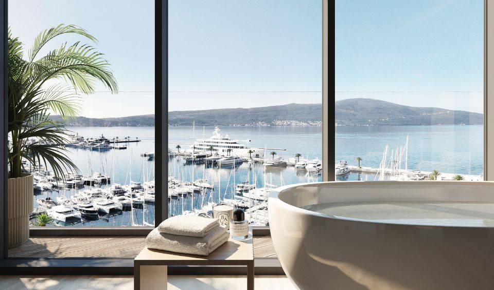 Elegant bathroom in Synchro Residences, Porto Montenegro, featuring a freestanding bathtub positioned near floor-to-ceiling windows that offer a stunning view of the marina and the Adriatic Sea, enhancing the tranquil atmosphere.