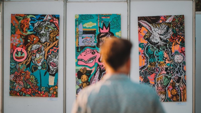 A person viewed from behind looking at three colorful, abstract paintings displayed on a white wall at Rubix Festival's Synchro Stage in Porto Montenegro. The artworks feature vibrant, chaotic designs with various characters, animals, and surreal elements.