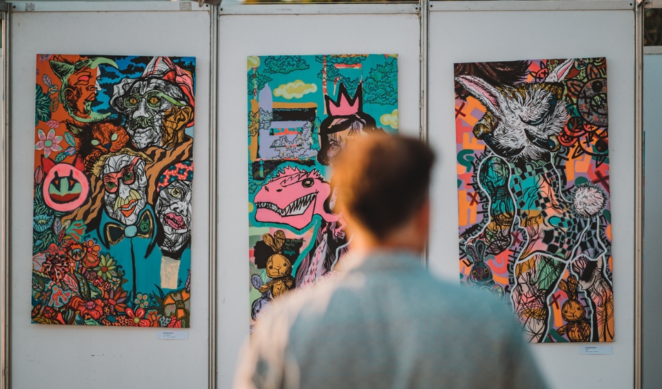 A person viewed from behind looking at three colorful, abstract paintings displayed on a white wall at Rubix Festival's Synchro Stage in Porto Montenegro. The artworks feature vibrant, chaotic designs with various characters, animals, and surreal elements.
