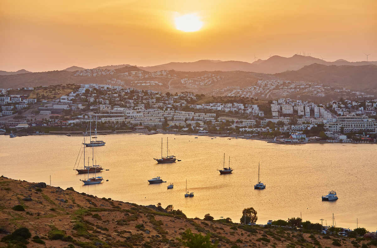 The sun sets over a tranquil bay dotted with yachts, with the silhouette of a hilly town and wind turbines on the horizon.