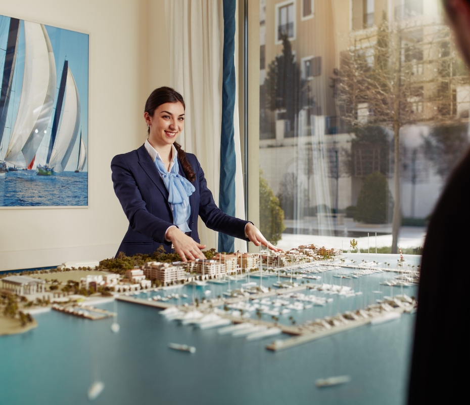 A businesswoman in a suit gestures at a detailed architectural model of Porto Montenegro marina, while discussing it with a man in the foreground. A large photograph of sailboats hangs on the wall behind her, symbolizing her successful career in the maritime industry.