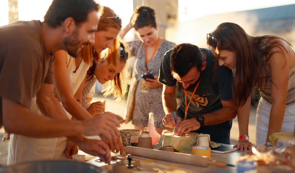 A group of people at Rubix Festival's Synchro Stage in Porto Montenegro leaning over a table, actively engaged in a hands-on craft activity under warm, natural light. Various tools and materials are scattered across the table, indicating a creative and collaborative project.