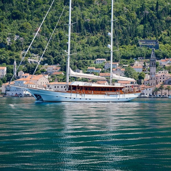 A classic sailing yacht with a wooden hull and white sails lowered, cruising in front of a picturesque coastal town.