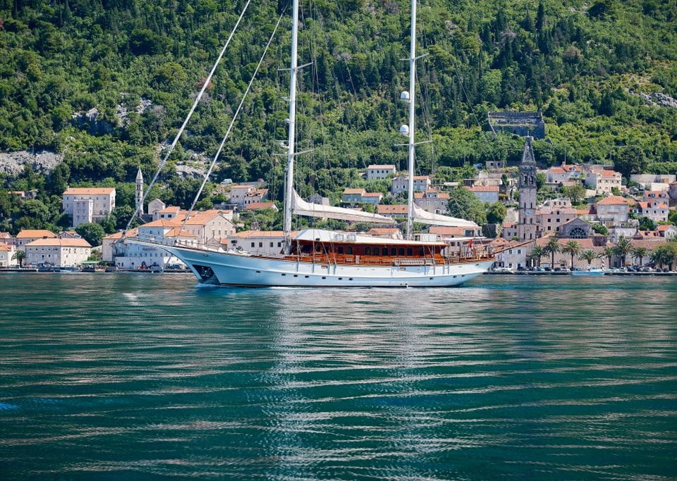 A classic sailing yacht with a wooden hull and white sails lowered, cruising in front of a picturesque coastal town.