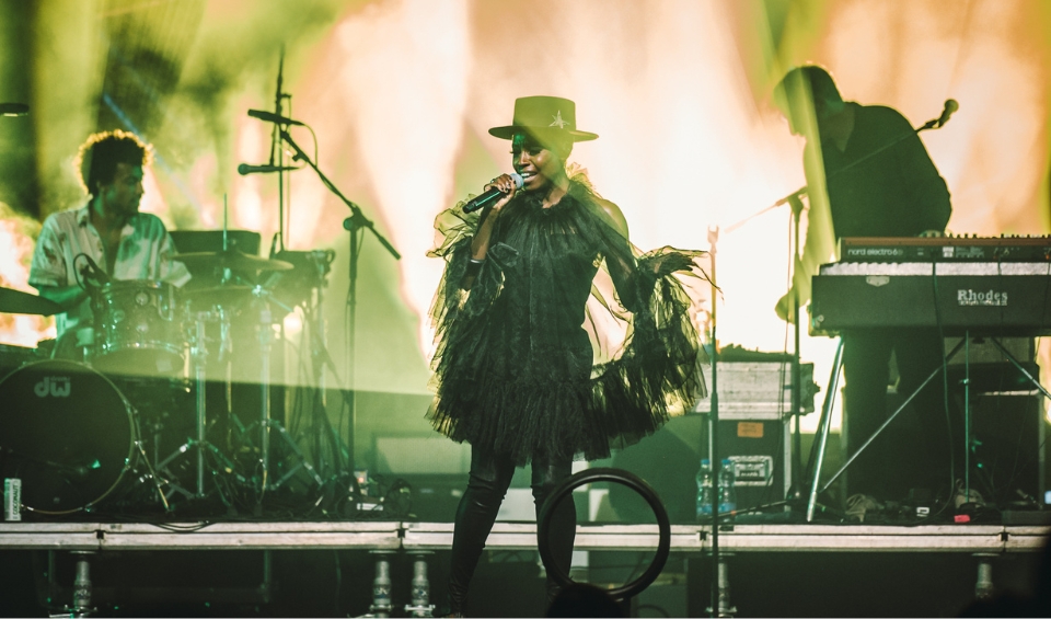 A dynamic concert scene at Rubix Festival's Synchro Stage in Porto Montenegro with a singer wearing a wide-brimmed hat and a flowing, dramatic outfit, performing under green and yellow lights. Behind the singer, a drummer and keyboardist play, adding to the vibrant and energetic atmosphere of the live performance.