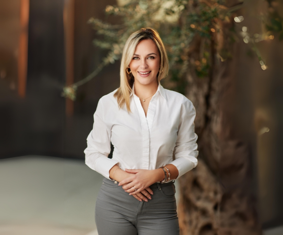 A friendly woman with blonde hair, wearing a white blouse and gray pants, smiles warmly while standing in the modern office, suggesting a welcoming environment.