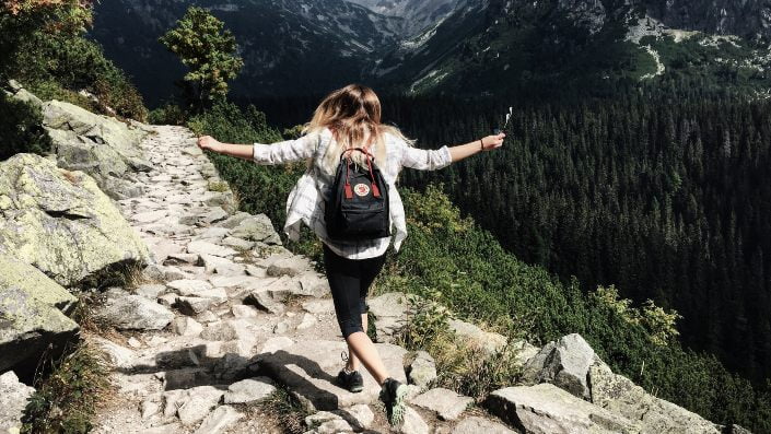A person with a backpack is joyfully hiking on a mountain trail surrounded by trees.