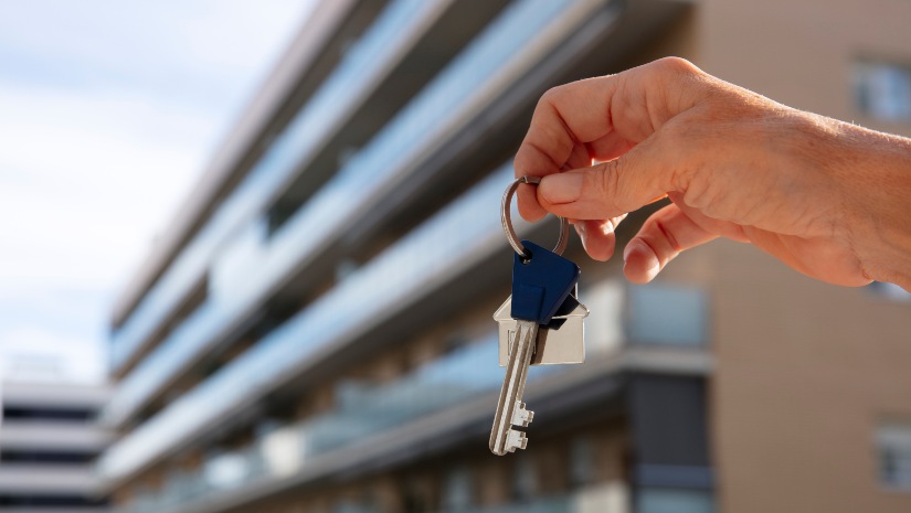 A hand holding keys in front of a modern apartment building in Porto Montenegro.