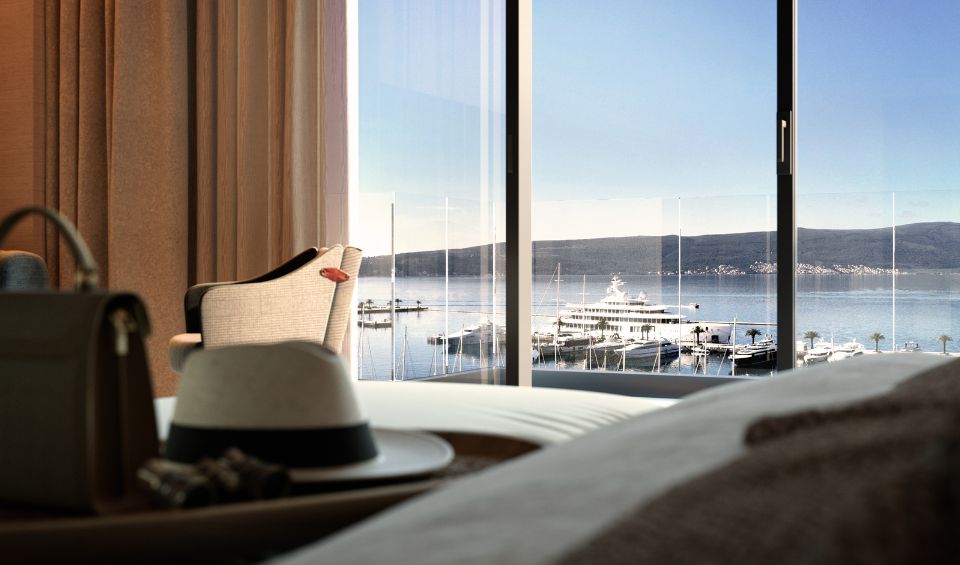 Room view from the IVO Residences at Porto Montenegro, highlighting a stylish armchair and a desk set against large panoramic windows that showcase a stunning marina and mountain backdrop.