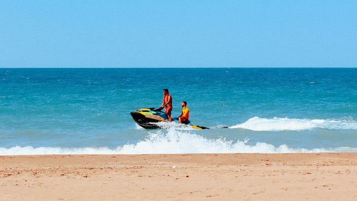 Two people are standing on a jet ski close to the shore of a blue sea, with a clear sky above and a sandy beach in the foreground.