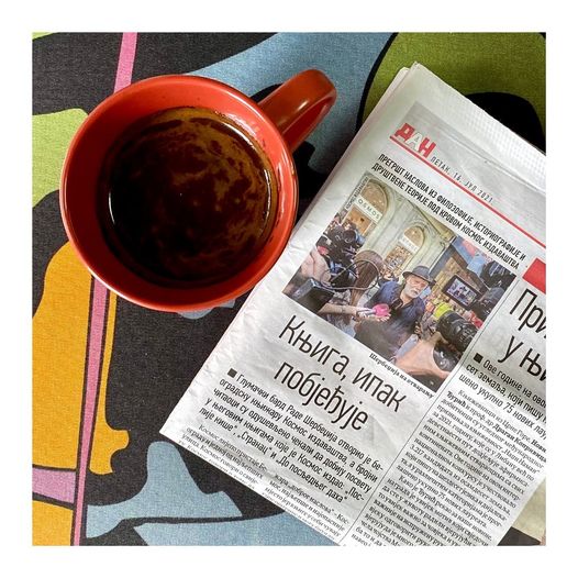 Kosmos Book picture featuring a newspaper with a Cyrillic headline, next to a red cup of coffee.