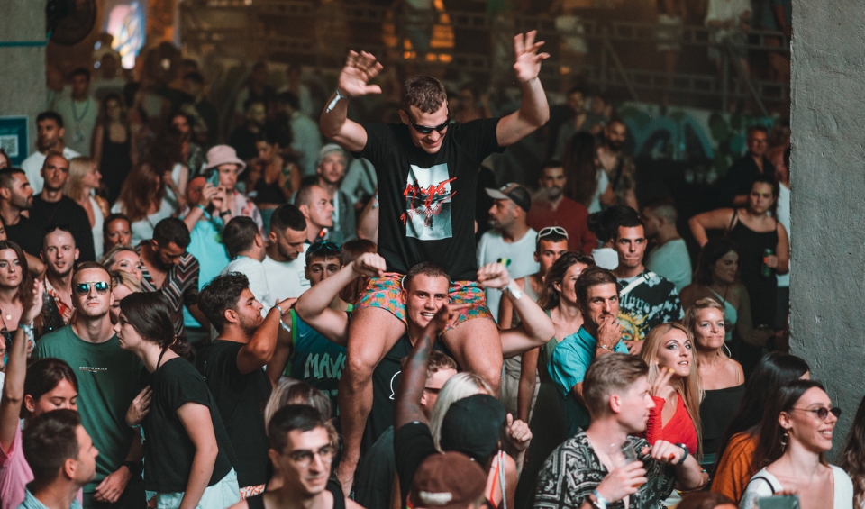 A lively crowd at Rubix Festival's Synchro Stage in Porto Montenegro, with one person sitting on another's shoulders, raising their hands in excitement. The audience, consisting of diverse individuals, appears engaged and cheerful, enjoying the vibrant atmosphere.