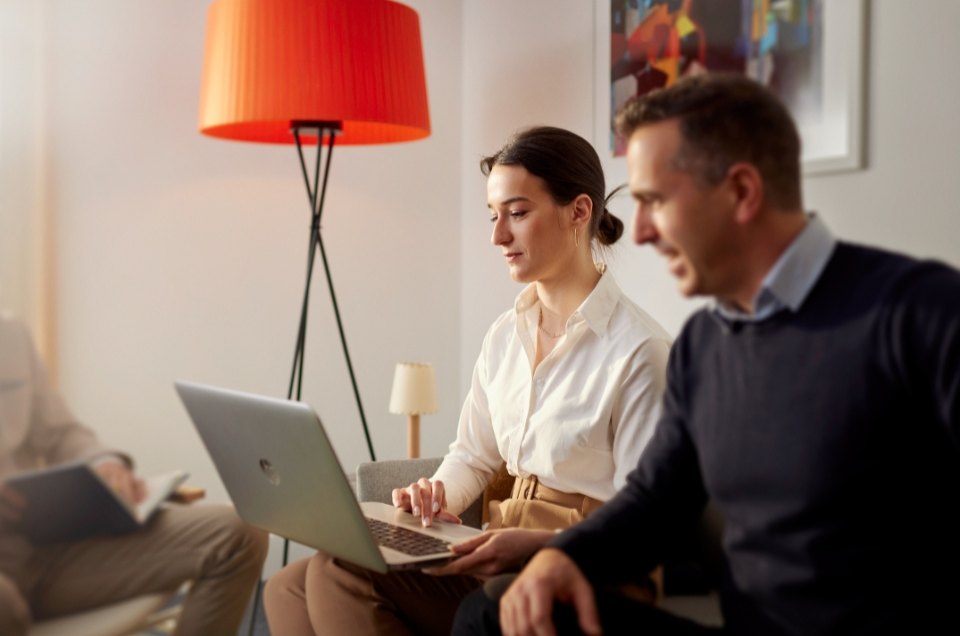 Two colleagues are in a modern office. The woman works on her laptop while the man discusses something. The room has an orange floor lamp and modern art.