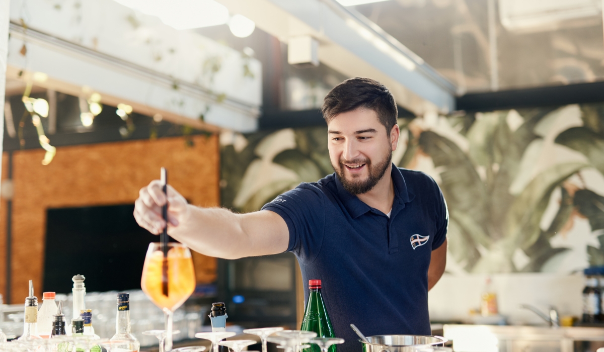 A smiling bartender in a navy polo shirt prepares a drink, creating a welcoming and engaging atmosphere behind the bar.