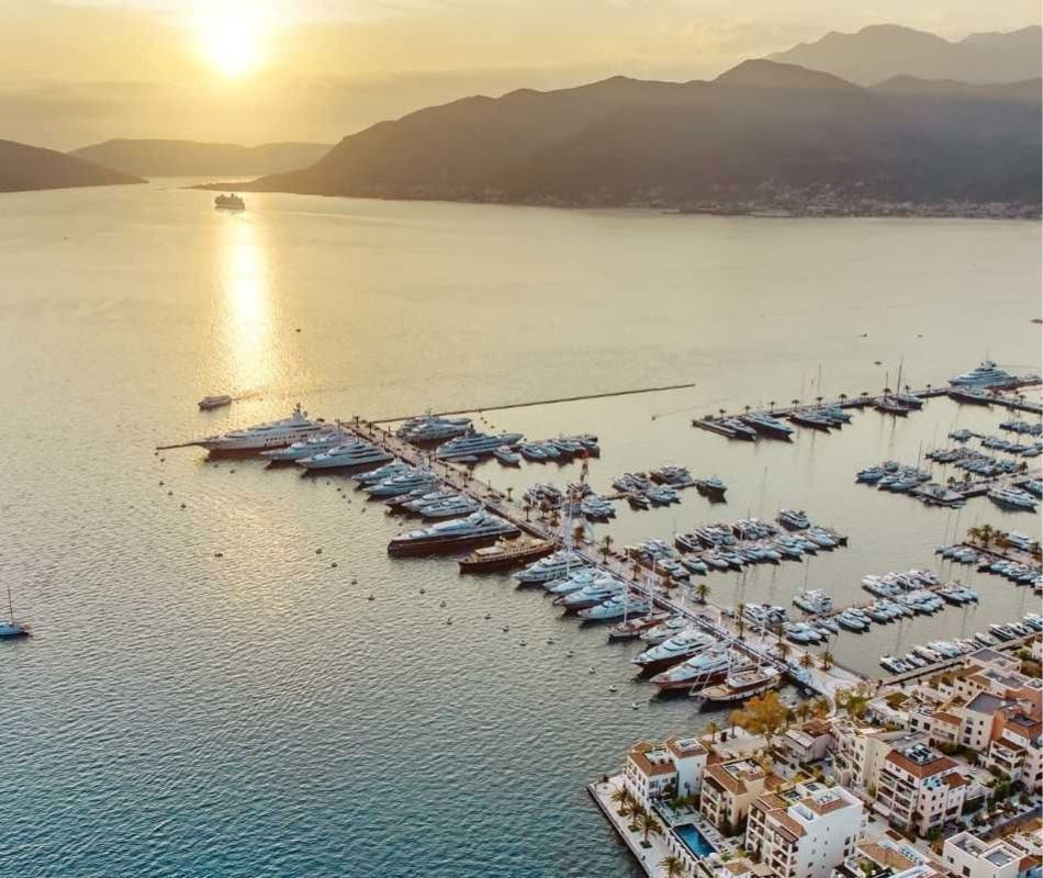 Aerial view of Porto Montenegro Marina at sunset, showcasing a vast array of docked yachts, surrounded by the serene waters of the Adriatic Sea and the mountainous landscape in the background.