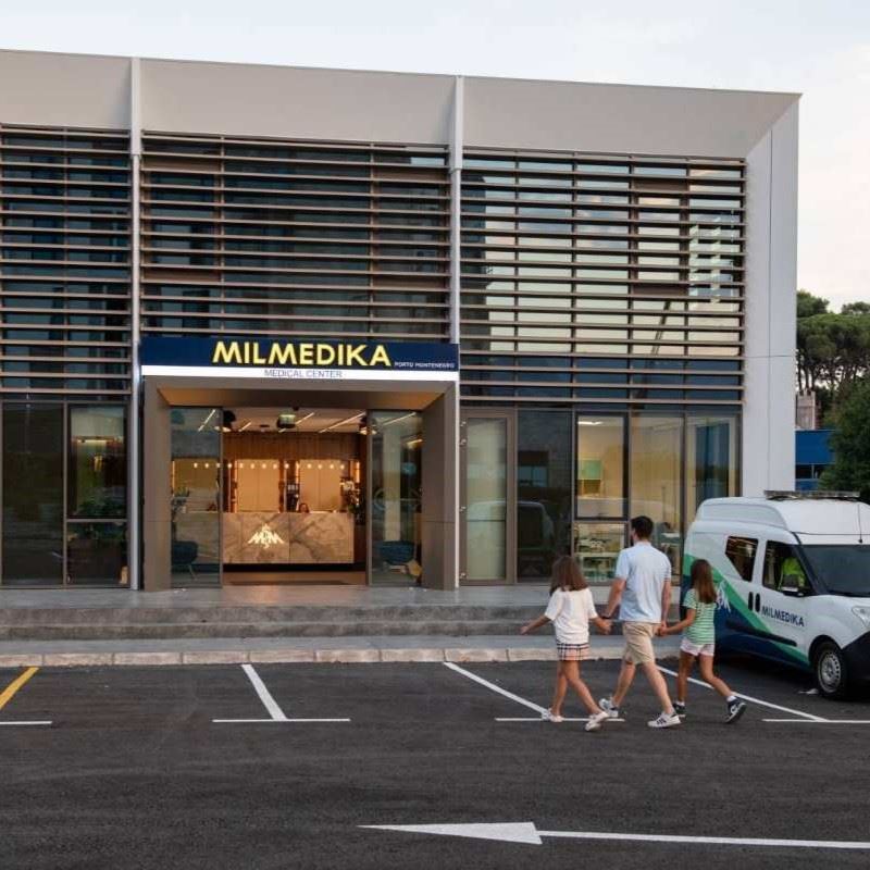 A modern medical center with a sign reading "MILMEDIKA Medical Center Porto Montenegro." The entrance has glass doors and a sleek interior with a marble reception desk. A man and two young girls walk towards the clinic, while a medical vehicle is parked nearby.
