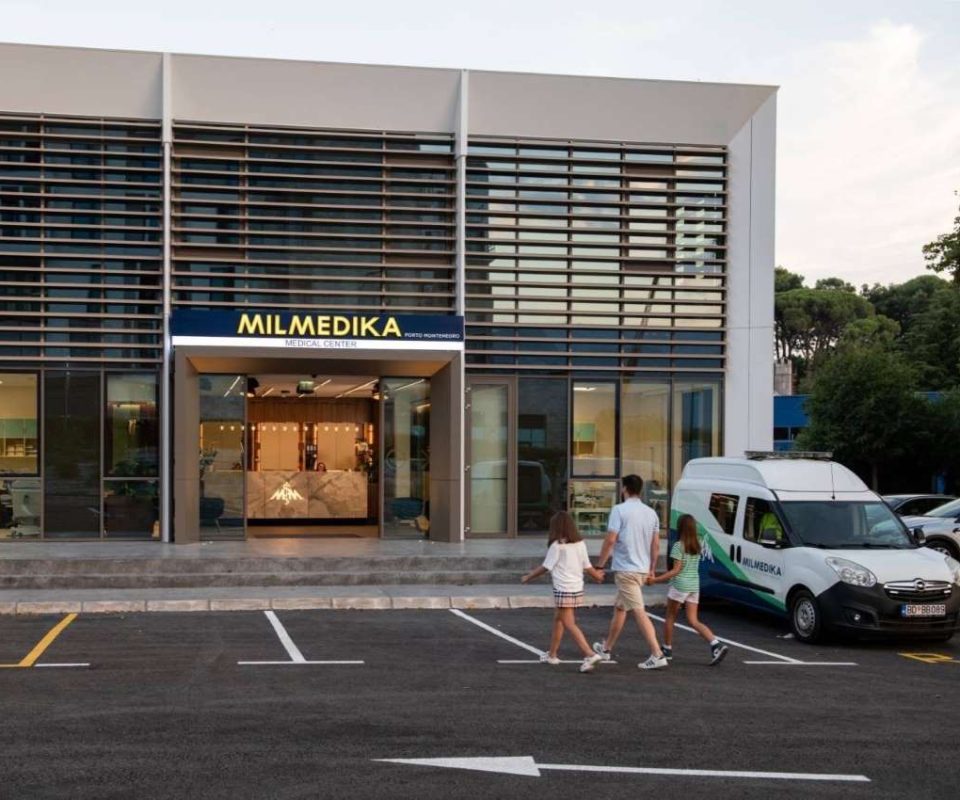 A modern medical center with a sign reading "MILMEDIKA Medical Center Porto Montenegro." The entrance has glass doors and a sleek interior with a marble reception desk. A man and two young girls walk towards the clinic, while a medical vehicle is parked nearby.