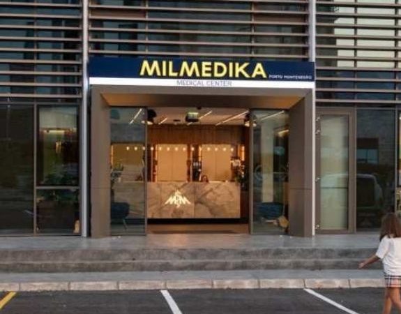 Front entrance of Milmedika Medical Center with a modern glass facade, featuring a large sign above the doorway. The clinic's reception desk is visible through the open doors, and a child is seen walking in the parking lot in front of the building.