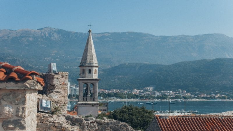 Scenic coastal town with a bell tower, historic architecture, boats along the shore, and mountains in the background.