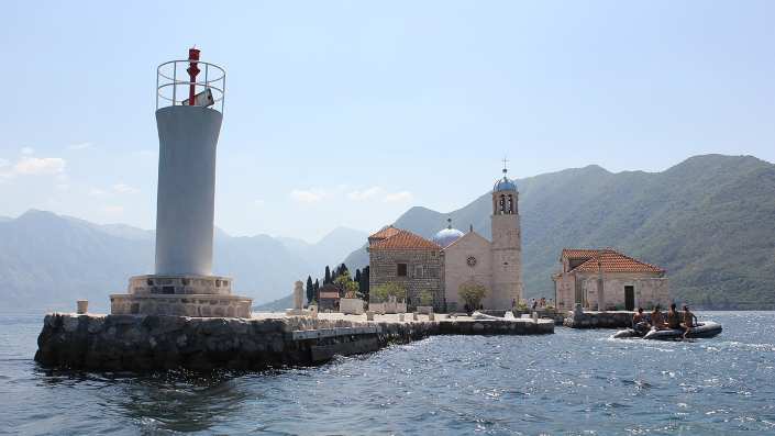 View of the island of Our Lady of the Rocks in Perast, with the iconic church and lighthouse situated on a small island in the middle of a sparkling blue bay.