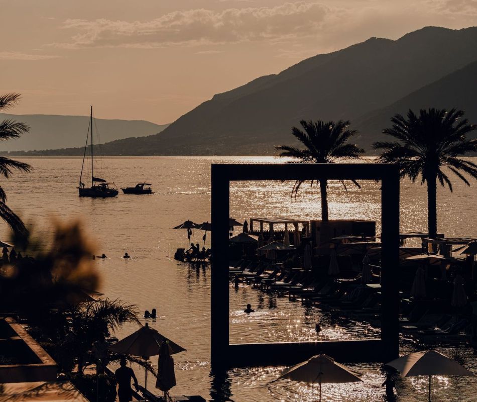 Sunset view at Porto Montenegro, capturing the serene atmosphere of the pool area with swimmers and sunbathers, silhouetted palm trees, and a tranquil bay reflecting the golden hues of the setting sun, framed by the picturesque mountain backdrop.