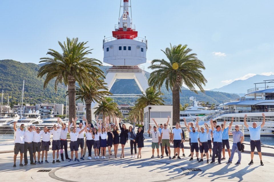 A team of marina employees posing together with raised hands, surrounded by palm trees, yachts, and a mountainous landscape on a sunny day.