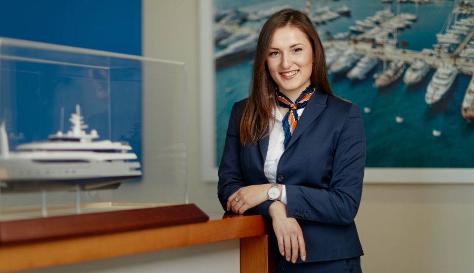 A friendly receptionist in a navy blazer stands at a desk with a model yacht, ready to assist, reflecting a welcoming and professional environment.