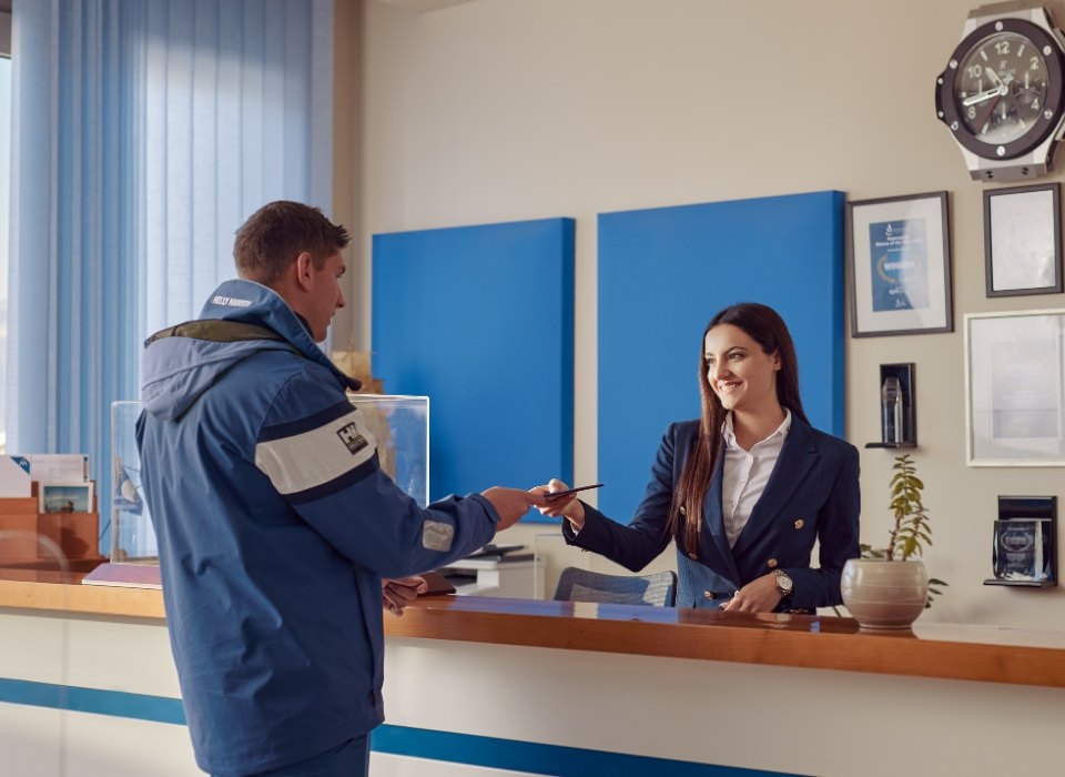 A friendly receptionist in a navy blazer hands a document to a man in a blue jacket, highlighting a professional and welcoming interaction.