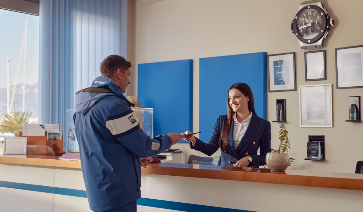 A friendly receptionist in a navy blazer hands a document to a man in a blue jacket, highlighting a professional and welcoming interaction.