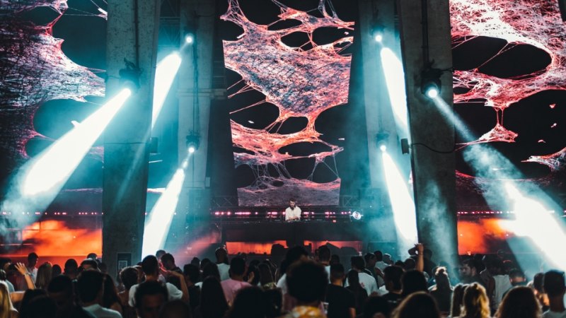 A crowded concert scene at Rubix Festival's Synchro Stage in Porto Montenegro, with a DJ performing on a stage surrounded by towering pillars and bright, white spotlights. The background features a large screen displaying abstract, web-like red and pink patterns.