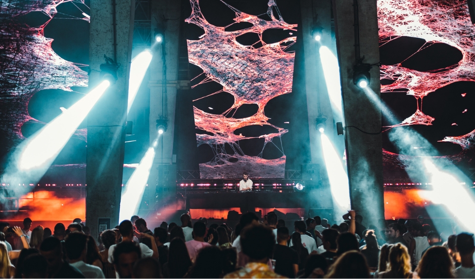 A crowded concert scene at Rubix Festival's Synchro Stage in Porto Montenegro, with a DJ performing on a stage surrounded by towering pillars and bright, white spotlights. The background features a large screen displaying abstract, web-like red and pink patterns.