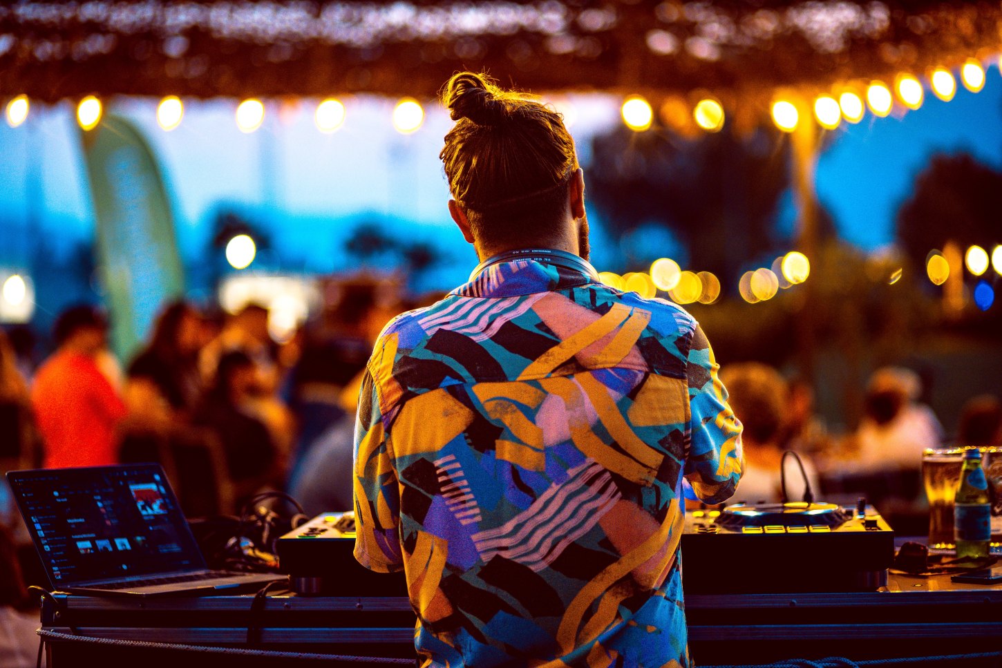 A DJ at the Rubix pre-party BBQ at Blue Room, Porto Montenegro, viewed from behind wearing a colorful, patterned shirt. The DJ is mixing music on a laptop and controller setup, with warm string lights and a lively crowd creating a festive atmosphere in the background.