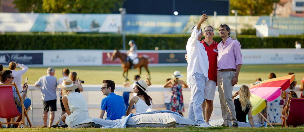 A group of spectators takes a selfie while watching a polo match, with other attendees relaxing on the grass and in lounge chairs. The scene is lively and vibrant, with players on horseback in the background, capturing the elegance and social atmosphere of a summer sporting event.
