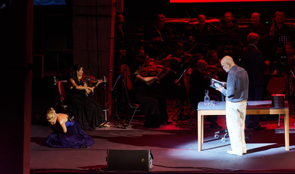A dramatic stage performance at Rubix Festival's Synchro Stage in Porto Montenegro featuring an orchestra in the background and a spotlight on John Malkovich reading from a book. To the left, a woman in a long dress is kneeling on the floor, while a violinist plays seated nearby, adding to the intense atmosphere of the scene.