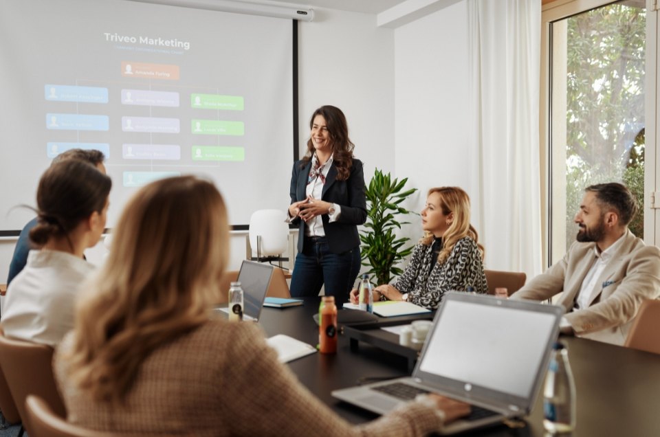 A woman in a black blazer presents to colleagues in a conference room. The presentation slide reads 'Triveo Marketing'