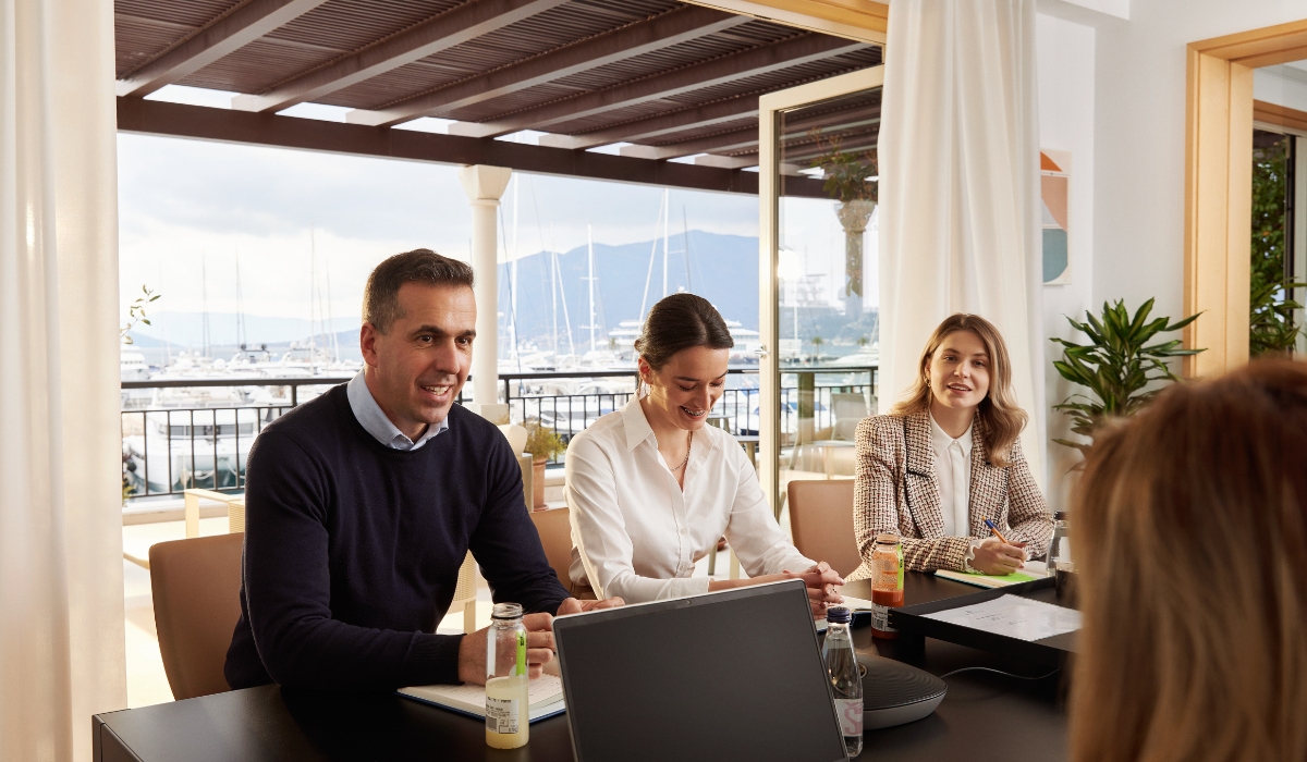 Three team members are in a meeting room with a marina view, engaged in a lively discussion, reflecting a collaborative work environment.