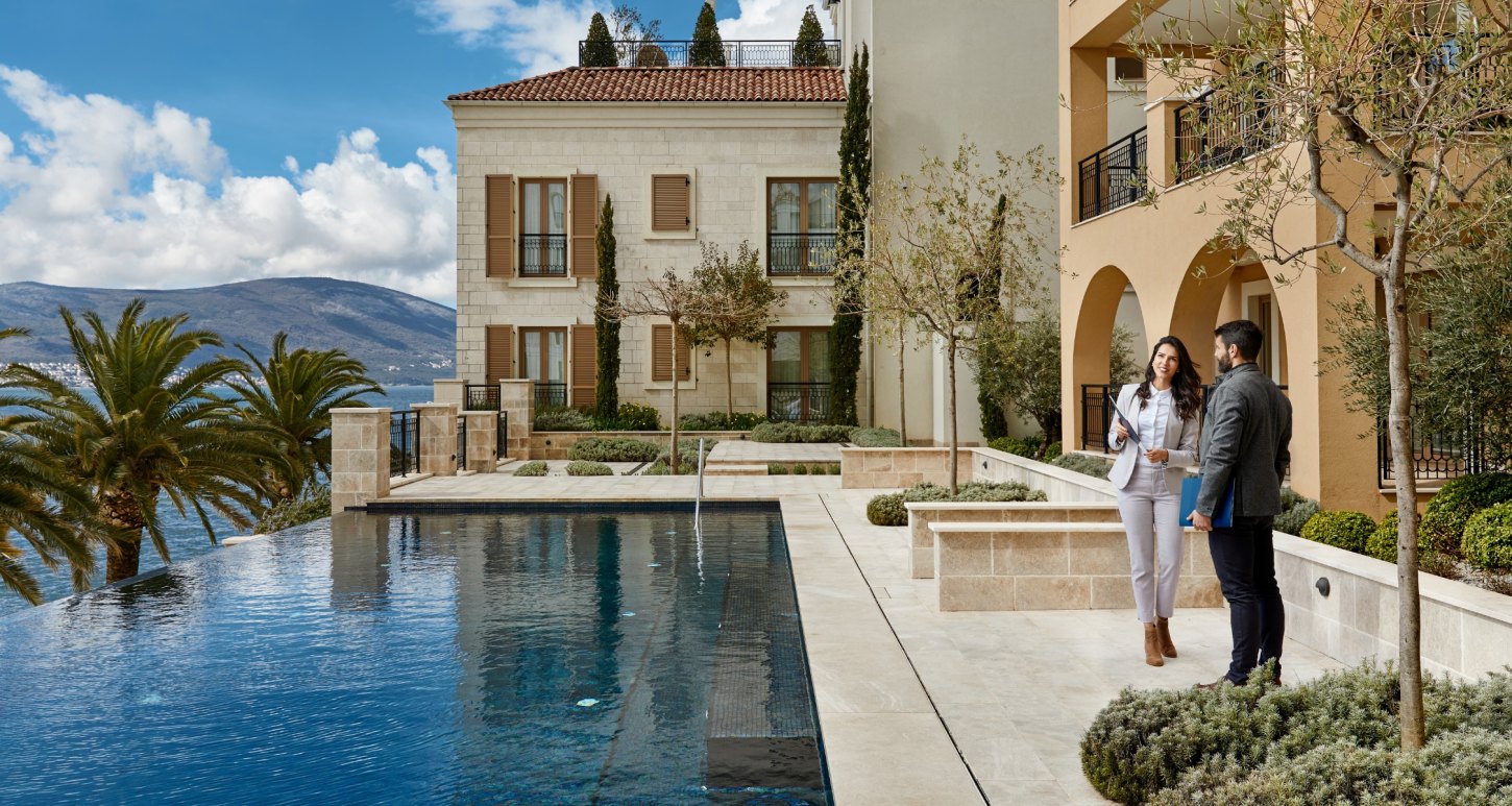 Two people having a conversation by a pool in front of a Mediterranean-style building with palm trees and mountains in the background, discussing career opportunities in the luxury village of Porto Montenegro.