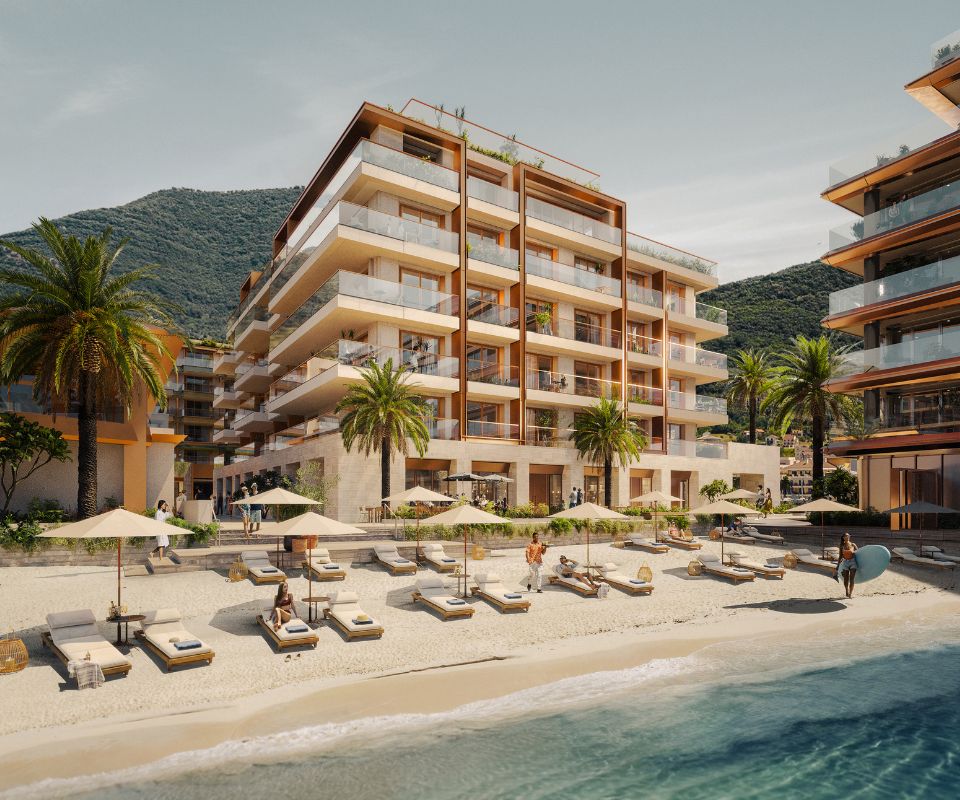 Daytime view of the IVO Residences in Porto Montenegro, a luxurious beachfront apartment complex with multiple balconies, palm trees, and sun loungers on a sandy beach.