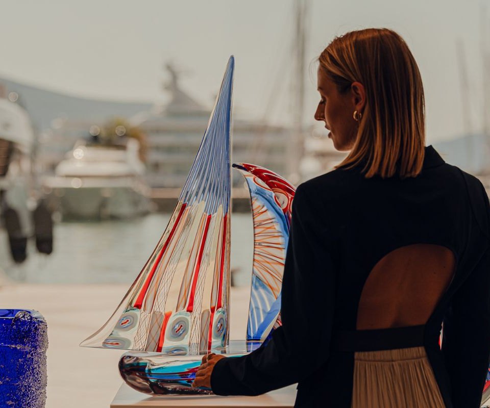 A woman at an art exhibition in Porto Montenegro, thoughtfully examining a colorful glass sculpture shaped like a sailboat, with luxurious yachts and the marina backdrop enhancing the artistic ambiance.